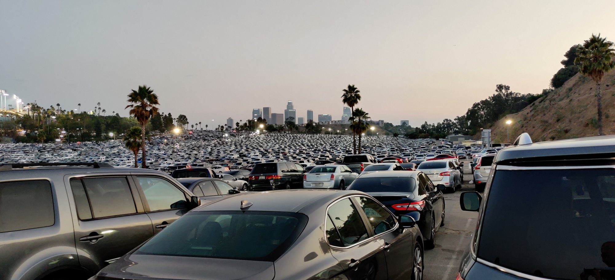 A vast, full parking lot at Dodger Stadium at dusk, thousands of white, grey, silver and black cars stretching toward the downtown Los Angeles skyline.