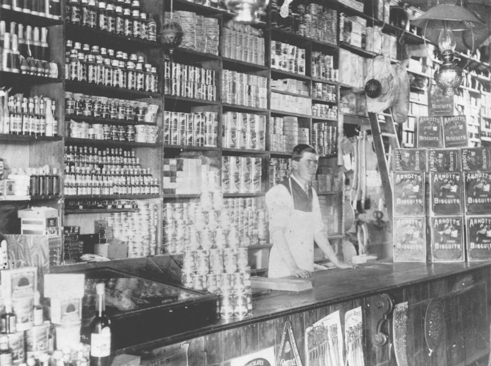 A black-and-white photograph of a 19th-century general store interior: a young grocer in an apron behind a wooden counter, shelves packed floor to ceiling with tins, bottles, and biscuit boxes.
