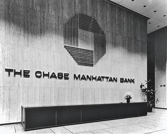 Black and white photo of a bank lobby with a large sculptural octagon logo above the words The Chase Manhattan Bank on a travertine wall.