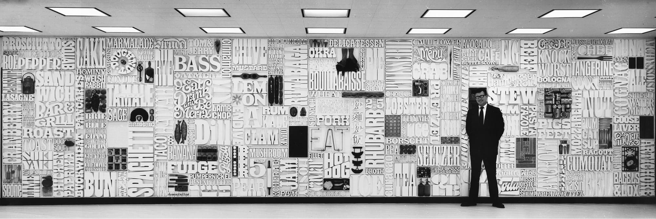 Black and white photo of Lou Dorfsman in a dark suit standing in front of the long CBS cafeteria wall, densely packed with carved wooden food words like SANDWICH, BASS, LOBSTER and SPAGHETTI.