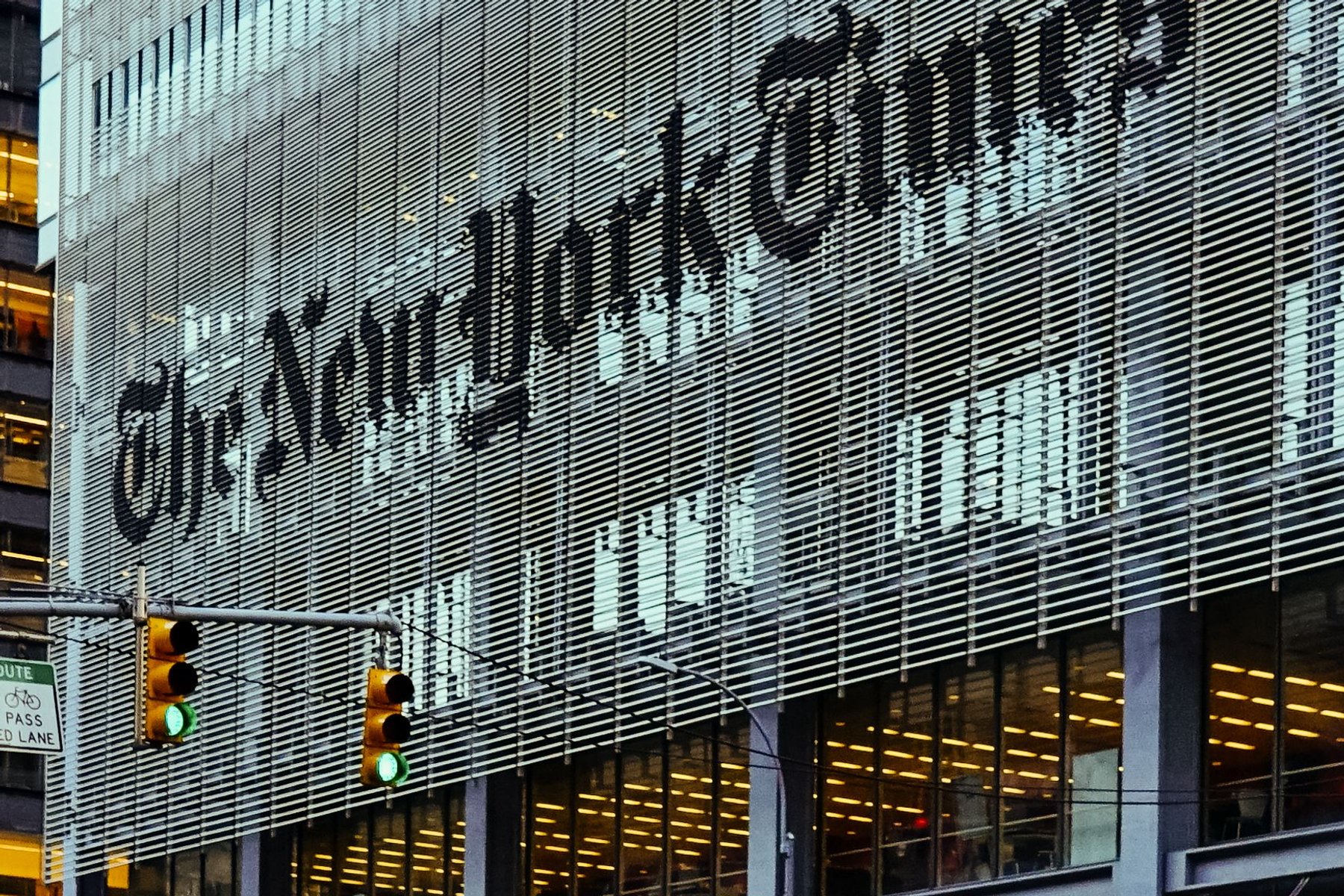 The New York Times logo in blackletter across the slatted facade of the Times building on Eighth Avenue, with traffic lights in the foreground.
