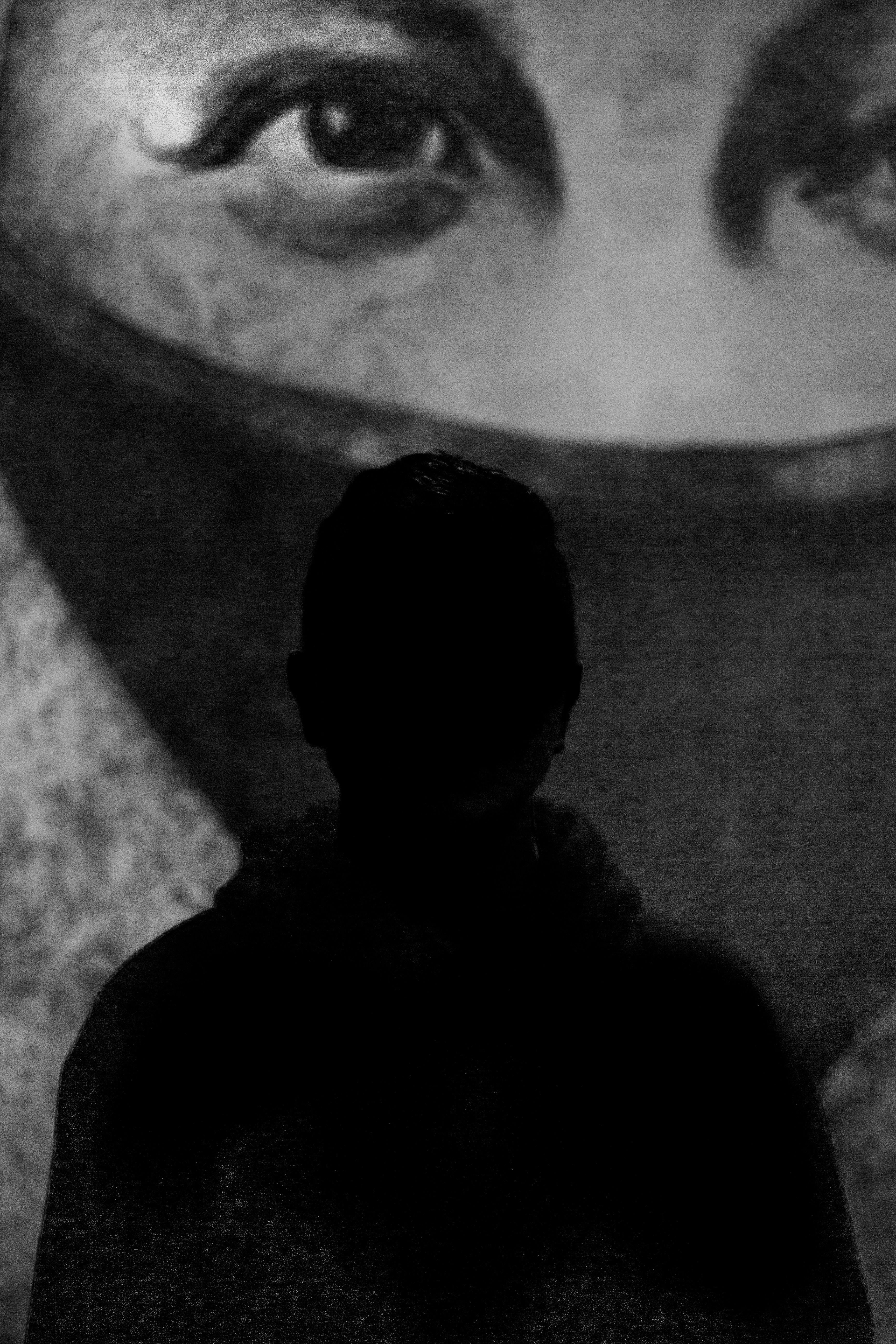 A black-and-white photograph of a silhouetted figure standing in front of a large projected close-up of a human face.