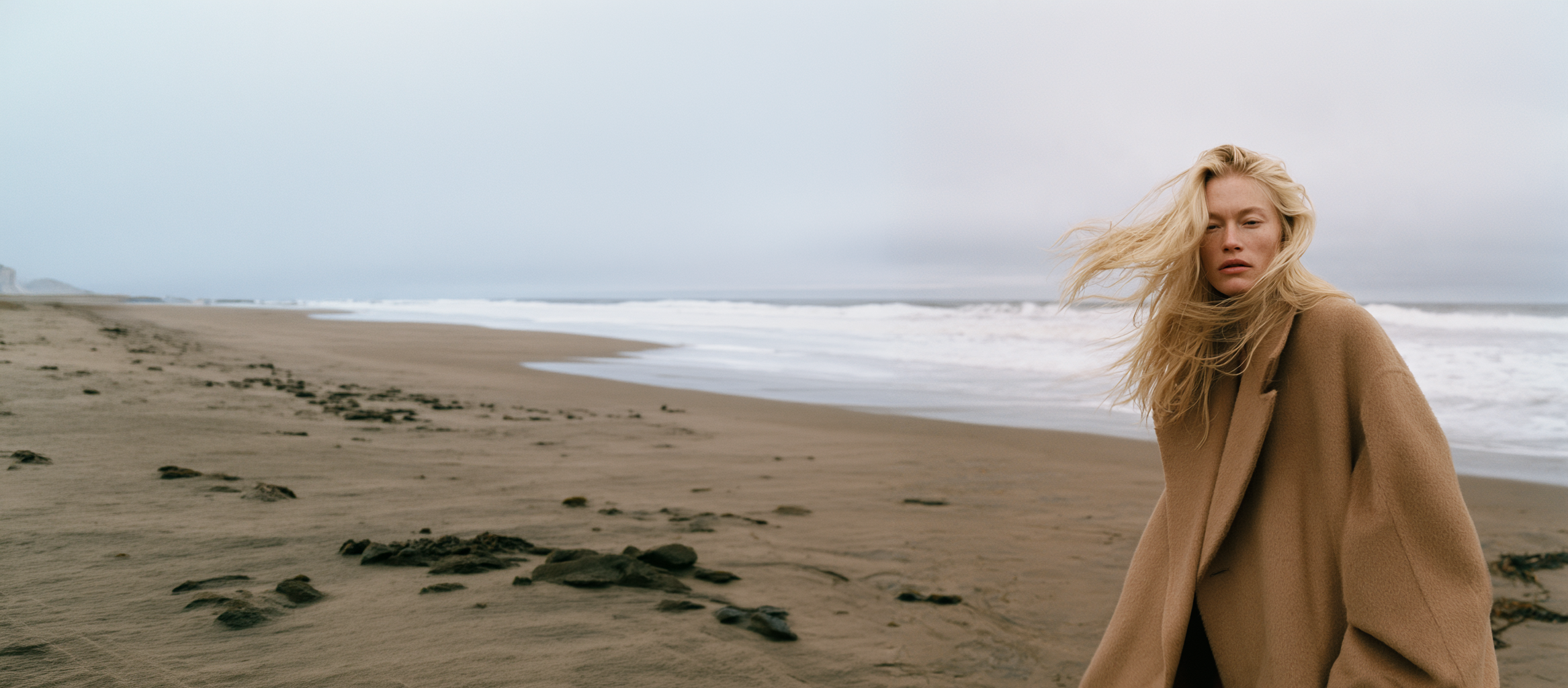 AI-generated fashion editorial: a blond model in an oversized camel wool coat, hair caught in the wind, on an overcast beach